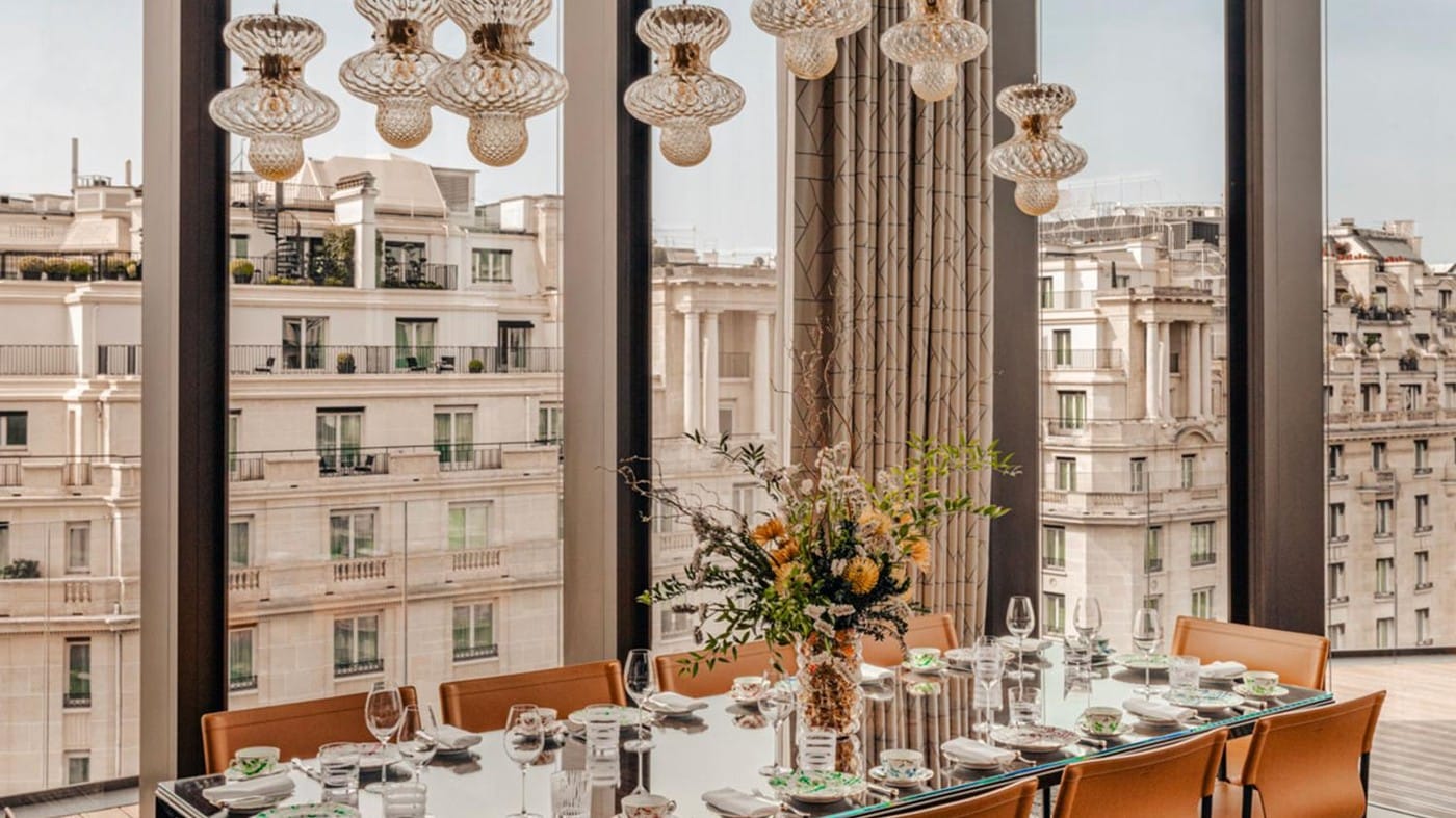 Une longue table dressée avec verres, porcelaine et un bouquet, sous une trentaine de lustres en verre suspendus, devant une baie vitrée ouverte sur les toits haussmanniens de Paris.