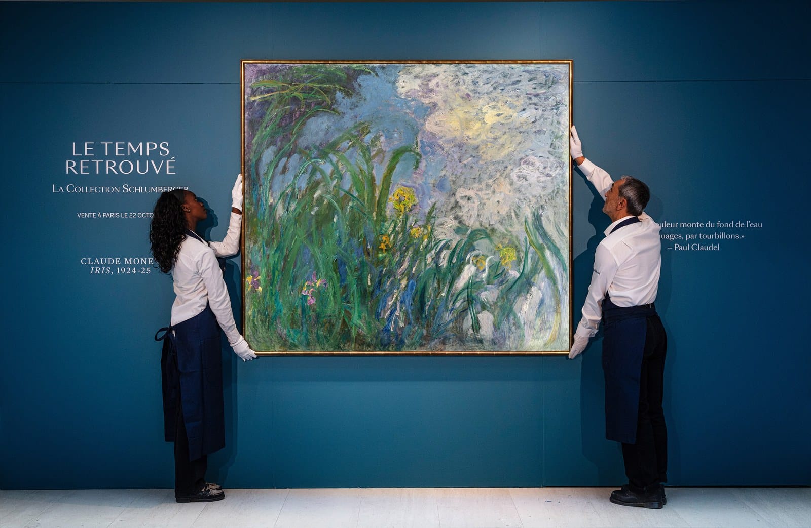 Deux manutentionnaires de Sotheby’s en gants blancs tiennent de chaque côté la toile Iris de Monet contre un mur bleu canard.