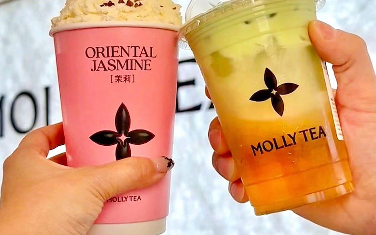 Deux mains tiennent des gobelets Molly Tea devant l’enseigne de la marque, le motif floral à quatre pétales bien visible sur le carton rose et sur le plastique.
