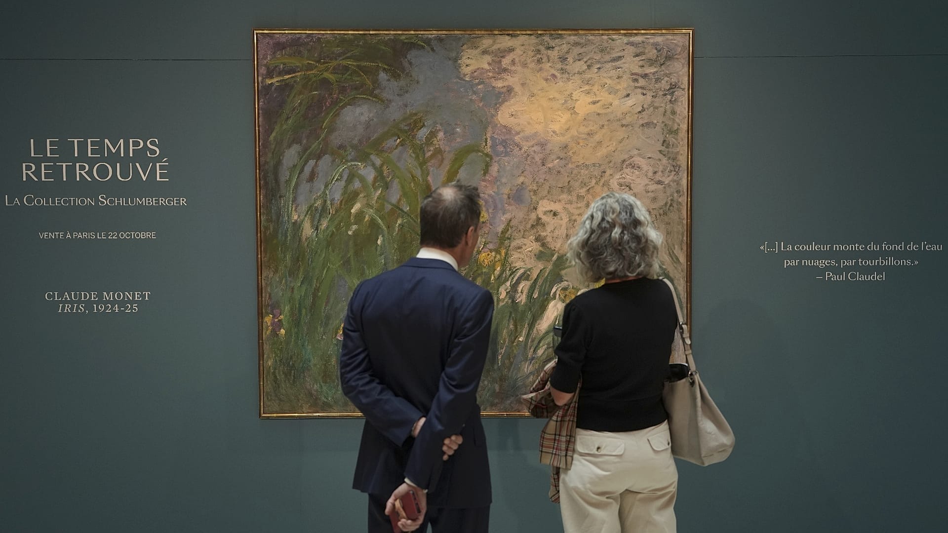 Un homme en costume et une femme aux cheveux gris, de dos, regardent la grande toile Iris de Claude Monet accrochée sur un mur vert sombre portant l’inscription Le Temps retrouvé, la collection Schlumberger.