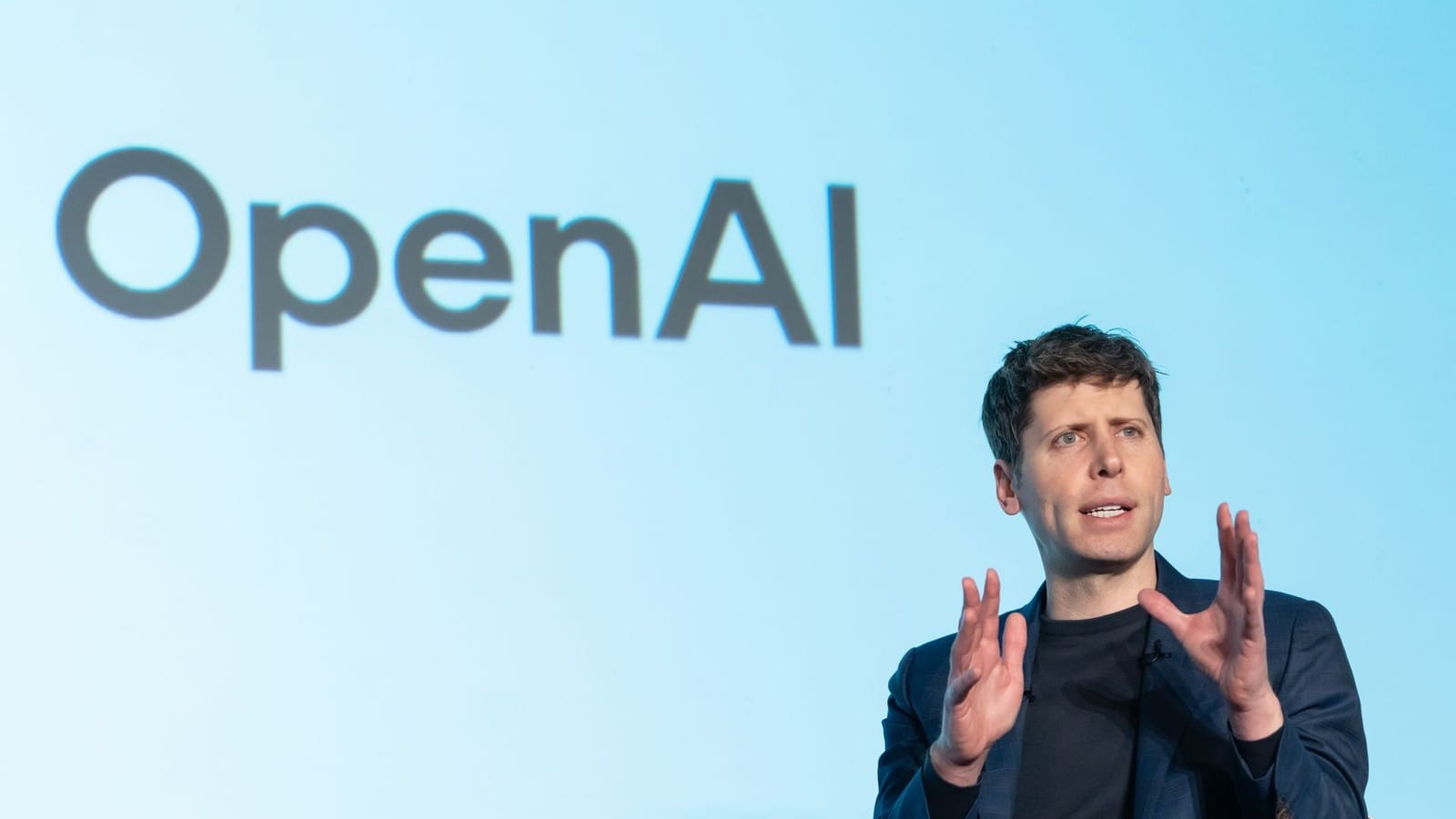 Sam Altman, assis, les deux mains levées en train de parler, devant un écran bleu clair affichant le logo OpenAI.