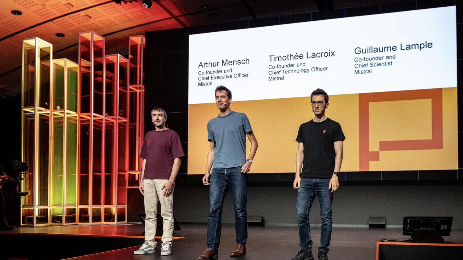 Les 3 cofondateurs de Mistral AI debout sur une scène, devant un écran affichant leurs noms et leurs fonctions, à côté d’une sculpture lumineuse aux tons orange et jaune.