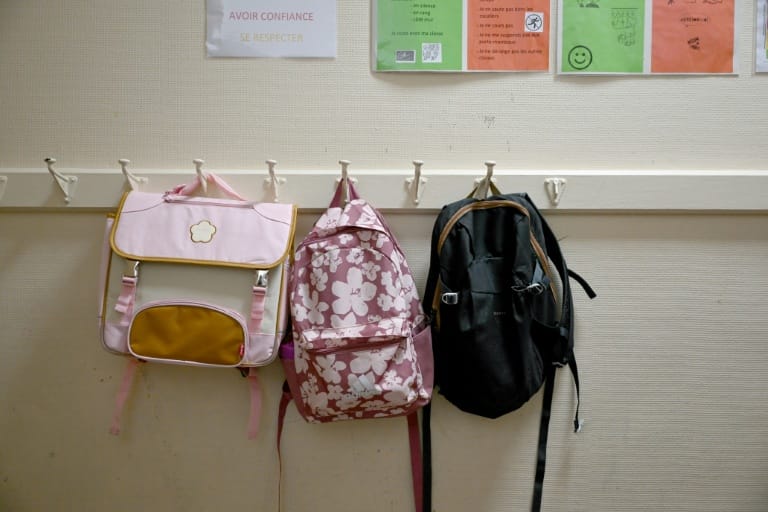 Trois cartables accrochés à des patères dans un vestiaire d’école, sous une affiche scolaire portant les mots « Avoir confiance, se respecter »