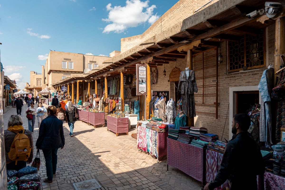 Ruelle marchande couverte de Toqi Telpak Furushon à Boukhara, étals de tissus et de robes, pancarte trilingue signalant un hammam du XVIe siècle, passants et touristes