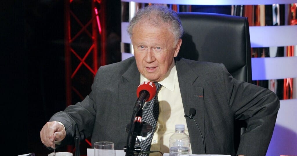 Philippe Bouvard assis en studio derrière un micro RTL, en veste et cravate, la bouche ouverte en pleine phrase