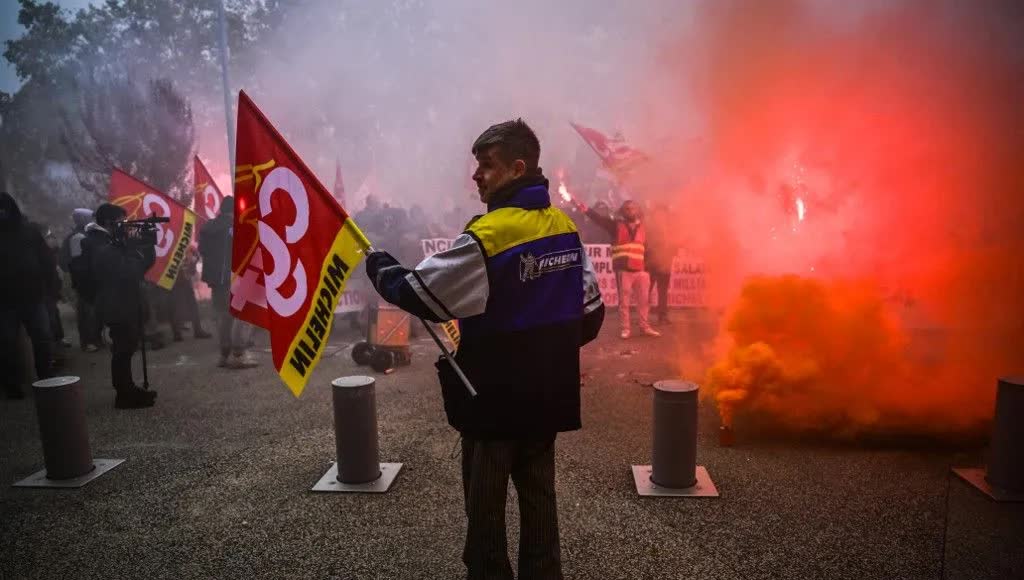 Un salarié en blouson Michelin, de dos et de profil, tient un drapeau CGT lors d’un rassemblement, au milieu d’une fumée rouge et d’autres manifestants avec banderoles.