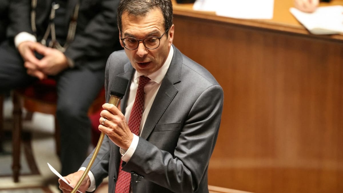 Jean-Pierre Farandou, costume gris et cravate rouge, parle dans un micro sur pied doré au milieu d’un hémicycle parlementaire, une feuille à la main