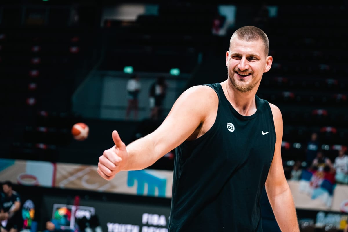 Nikola Jokic, en débardeur noir sur le parquet d’une salle, souriant et levant le pouce vers le côté, gradins et ballon flous en arrière plan.