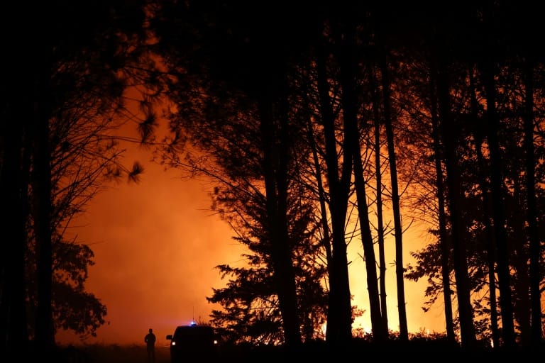 Silhouette d’un pompier et d’un véhicule de secours devant un rideau de pins noirs, sur un ciel entièrement orange saturé par les flammes et la fumée, de nuit, près de Luglon dans les Landes