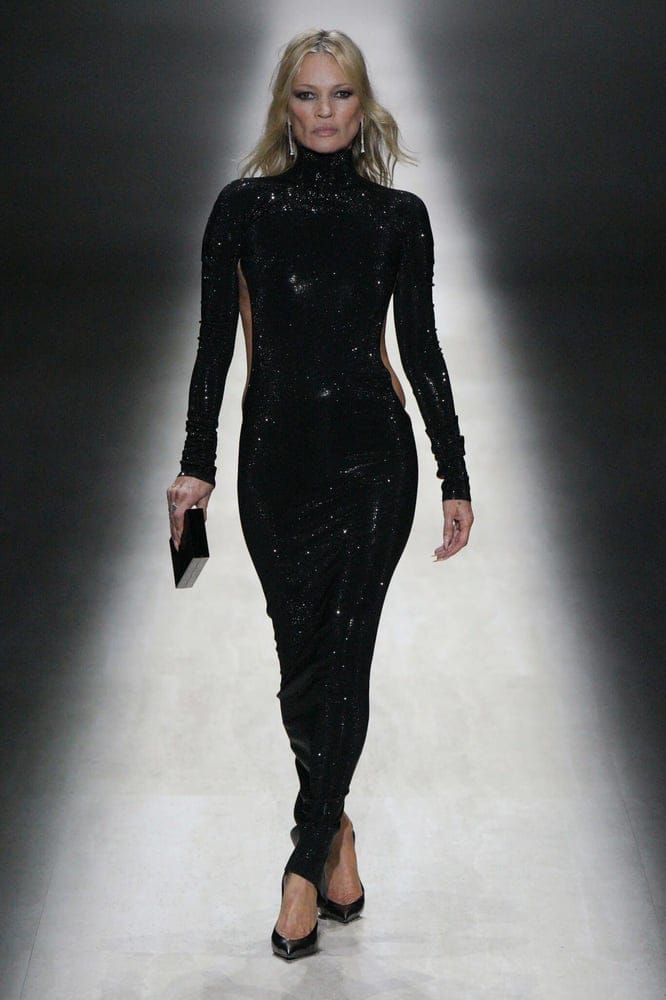 Kate Moss avance seule sur un podium de défilé, en robe longue noire à sequins dos nu, une pochette à la main, visage de face.