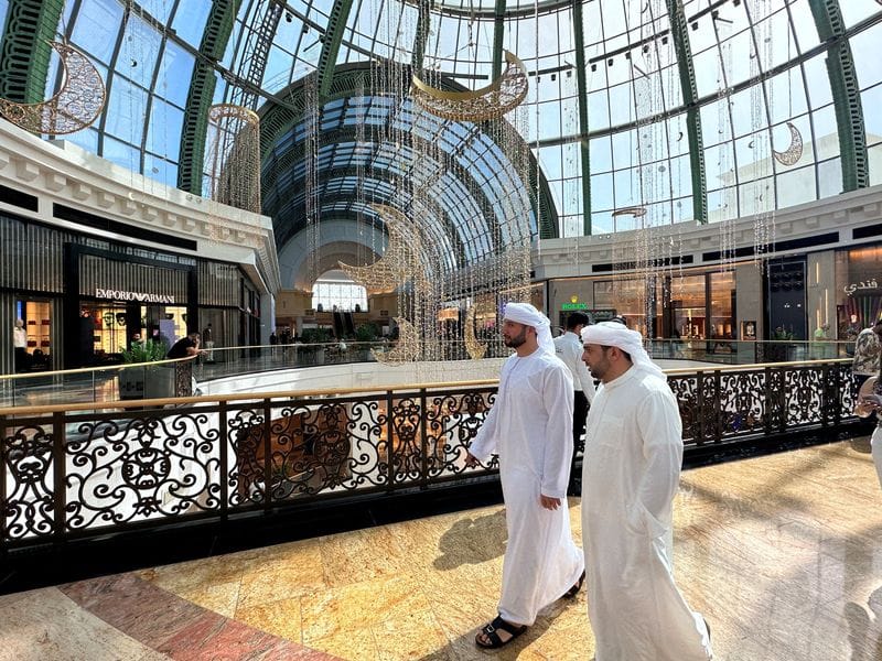 Deux hommes en kandura blanche marchent dans la galerie du Mall of the Emirates, devant les boutiques Emporio Armani et Boss, sous la verrière et des décorations de croissants dorés.