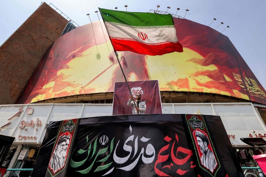 Un homme brandit un grand drapeau iranien sur une estrade, devant une façade couverte de banderoles et de portraits, dans une rue commerçante