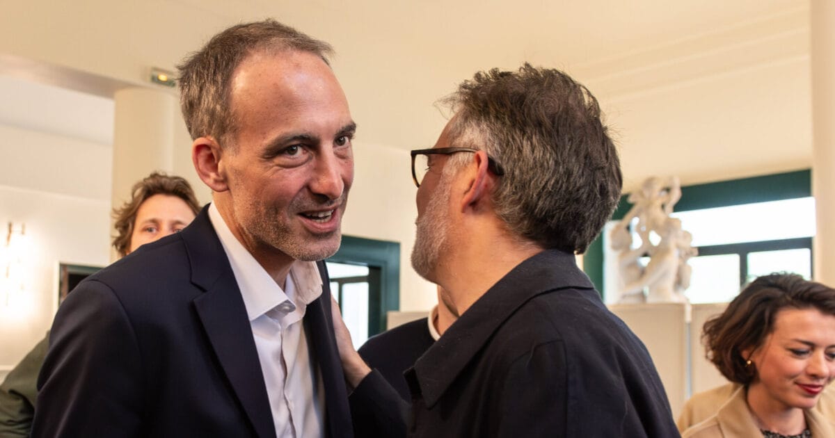 Raphaël Glucksmann, en costume sombre, sourit en conversation rapprochée avec un homme à lunettes qui lui tient l’épaule, entouré d’autres participants.