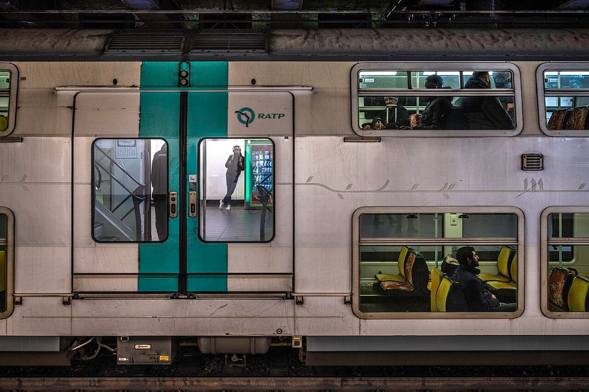 Une rame RER A à deux niveaux exploitée par la RATP, arrêtée aux quais souterrains de la gare d’Auber à Paris. Des voyageurs assis derrière les vitres, un voyageur debout dans l’encadrement 