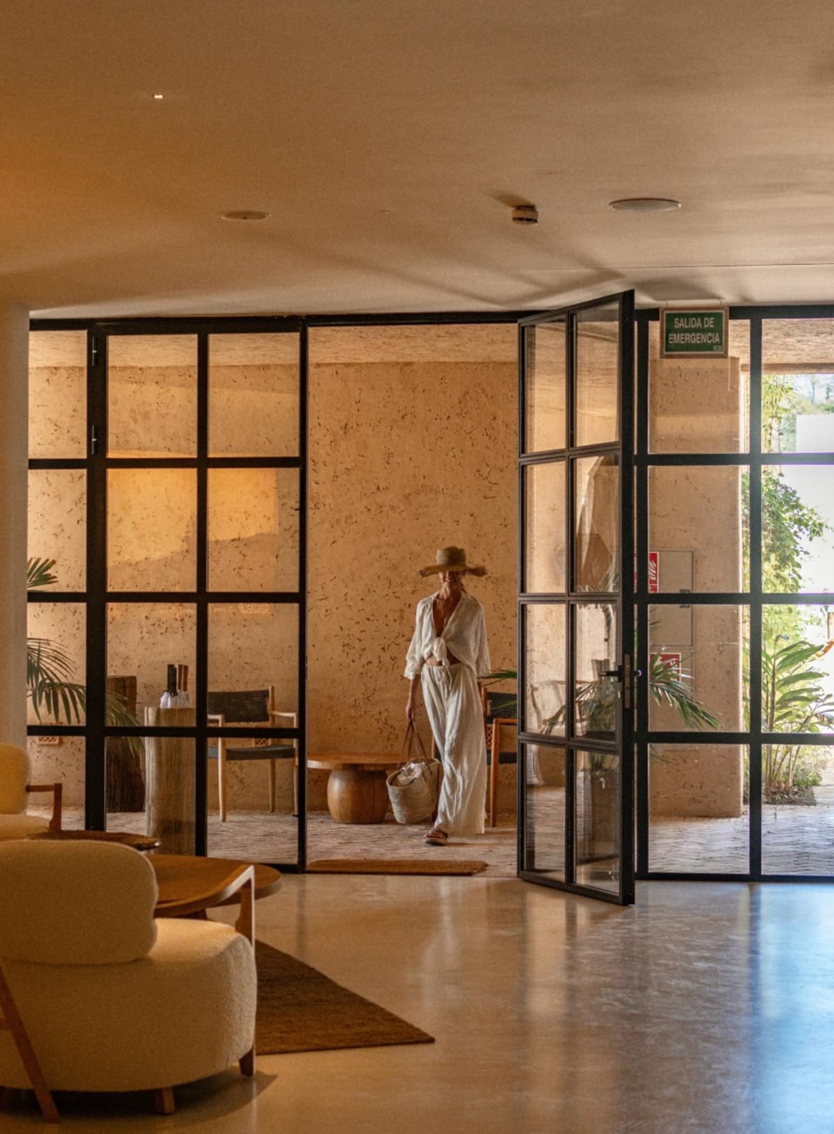 Intérieur d’un centre de longévité méditerranéen, portes vitrées ouvertes sur un patio, une cliente traverse la pièce en tenue de lin clair.