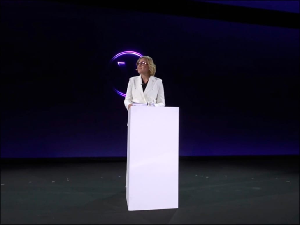 Henna Virkkunen, vice présidente exécutive de la Commission européenne, debout à un pupitre blanc sur une scène sombre éclairée par un projecteur, un anneau lumineux violet en fond d’écran.