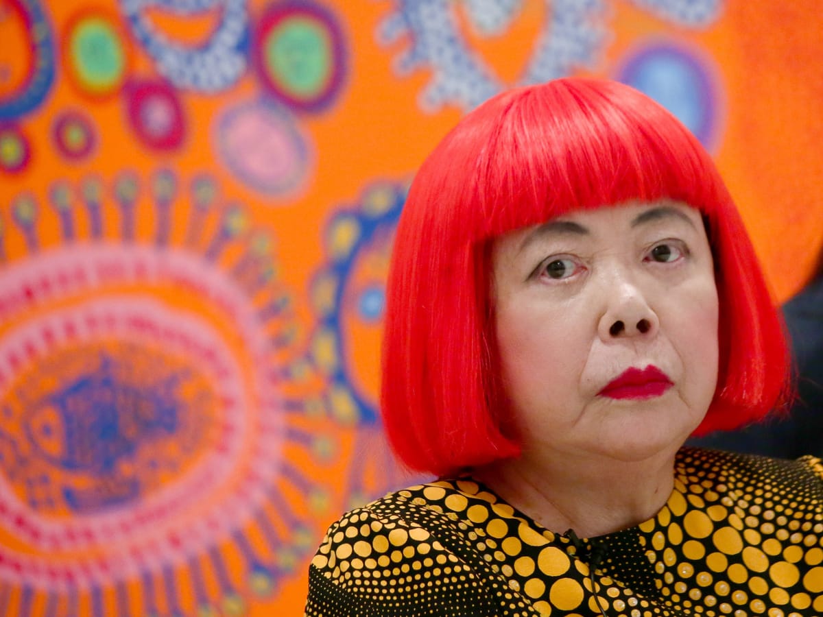 Yayoi Kusama, perruque rouge vif à frange, robe noire à pois jaunes, regardant hors champ devant une de ses toiles orange couverte de motifs colorés.