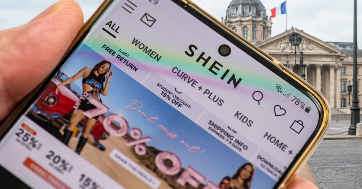 Une main tient un smartphone affichant l’application Shein et ses bandeaux de remises, devant la façade à colonnes d’un bâtiment officiel français où flotte le drapeau tricolore.